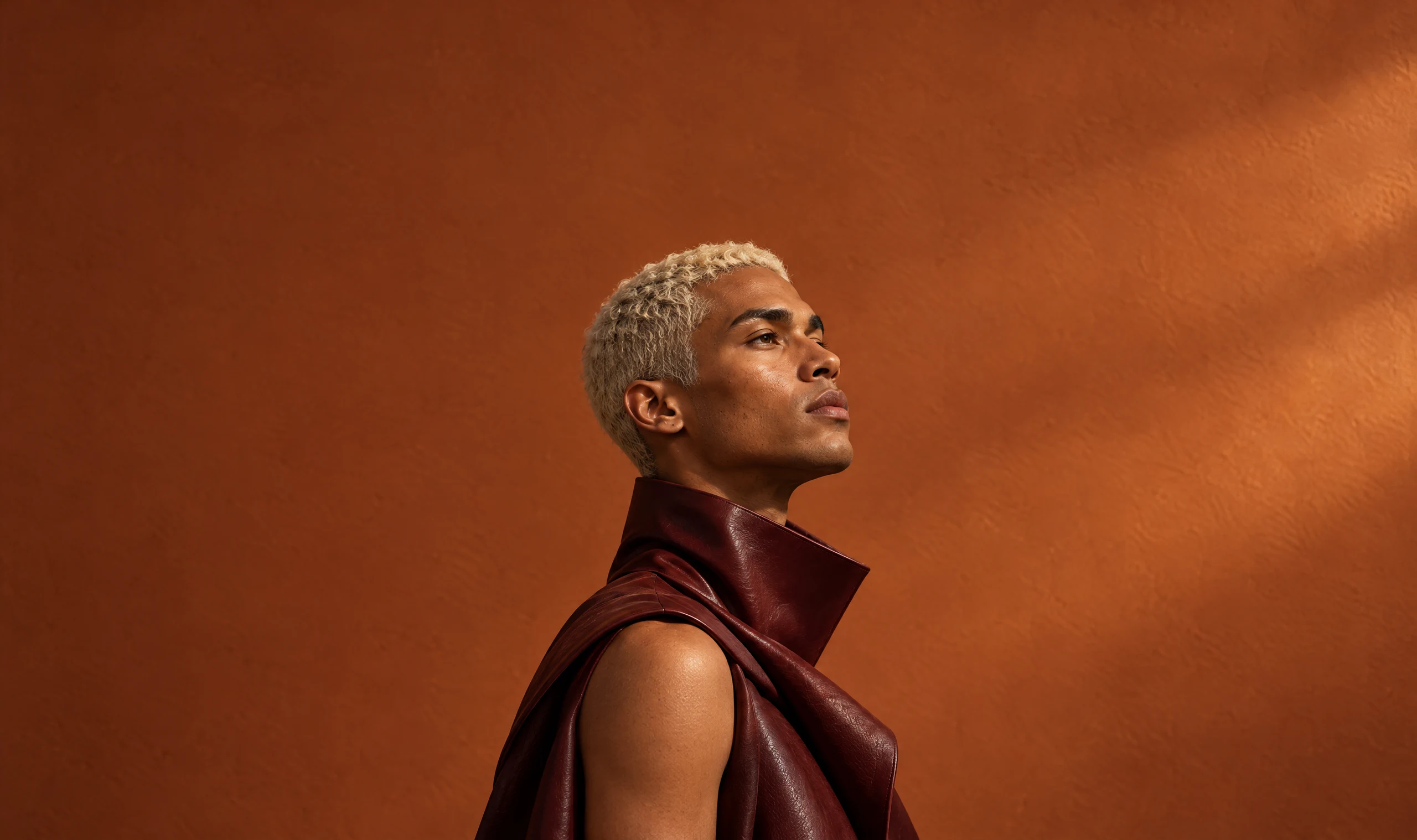Profile portrait of a man with bleached platinum hair in a sculptural oxblood leather collar, looking up against a sunlit terracotta wall